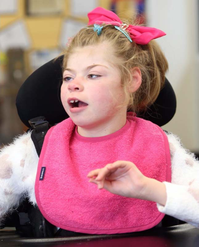 Girl in wheelchair wears a pink bib and matching pink bow in her hair