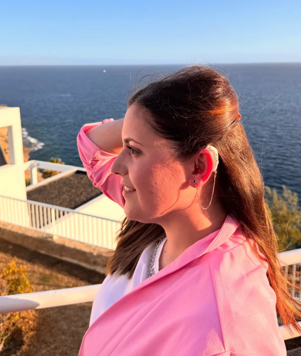 Woman wears a pink jacket and pulls back her hair to show a hearing aid cover with a chain to an earring